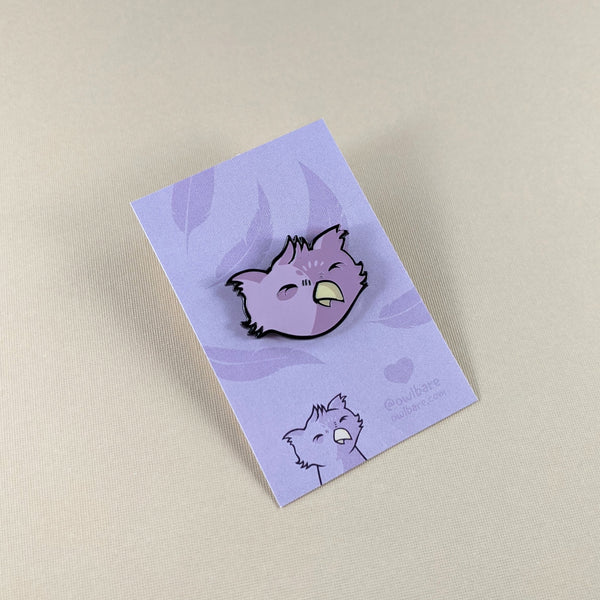 The owlbare enamel pin depicts the head of a purple, fuzzy owlbare. His eyes are closed because he's smiling and he has a tiny yellow beak and tufty ears. The pin has one post and comes with a black rubber clutch and purple backing card with a feather motif.