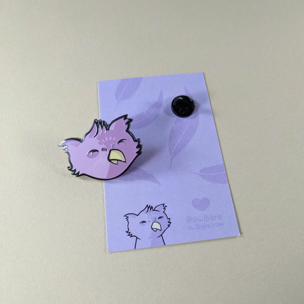 The owlbare enamel pin depicts the head of a purple, fuzzy owlbare. His eyes are closed because he's smiling and he has a tiny yellow beak and tufty ears. The pin has one post and comes with a black rubber clutch and purple backing card with a feather motif.