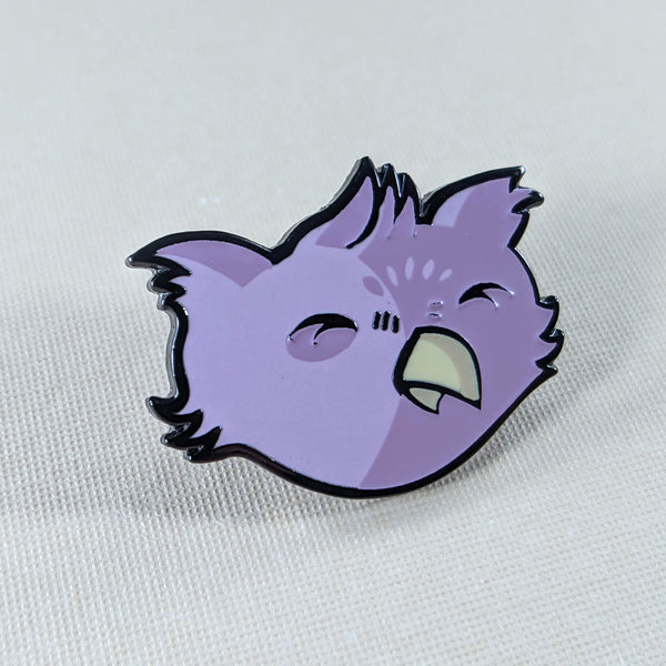 The owlbare enamel pin depicts the head of a purple, fuzzy owlbare. His eyes are closed because he's smiling and he has a tiny yellow beak and tufty ears. The pin has one post and comes with a black rubber clutch and purple backing card with a feather motif.