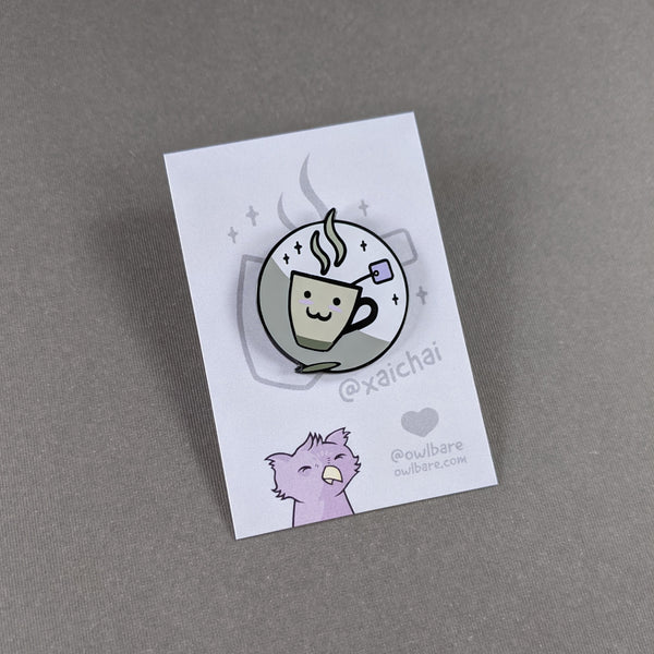 The xaichai enamel pin depicts a cute coffee mug with chai teabag hanging out. The mug is hella kawaii and smiling with rosey cheeks. The pin has one post and comes with a black rubber clutch and purple backing card.