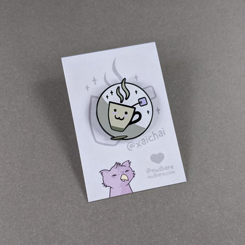 The xaichai enamel pin depicts a cute coffee mug with chai teabag hanging out. The mug is hella kawaii and smiling with rosey cheeks. The pin has one post and comes with a black rubber clutch and purple backing card.