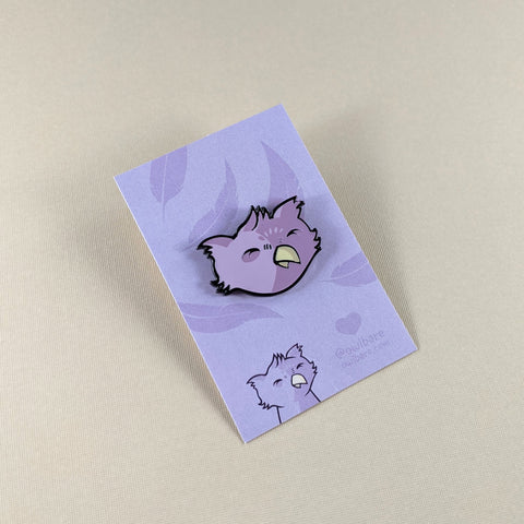 The owlbare enamel pin depicts the head of a purple, fuzzy owlbare. His eyes are closed because he's smiling and he has a tiny yellow beak and tufty ears. The pin has one post and comes with a black rubber clutch and purple backing card with a feather motif.