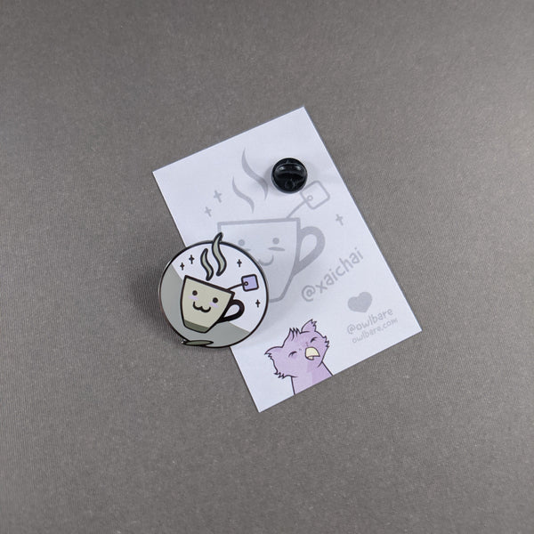 The xaichai enamel pin depicts a cute coffee mug with chai teabag hanging out. The mug is hella kawaii and smiling with rosey cheeks. The pin has one post and comes with a black rubber clutch and purple backing card.